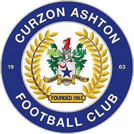 Curzon Ashton Football Club crest featuring a blue circular design with a green coat of arms, surrounded by golden laurel leaves. Founded in 1963.