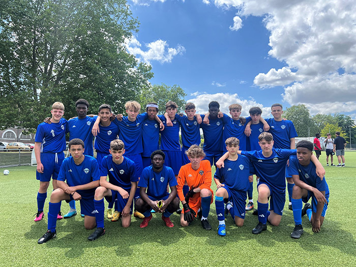 Youth soccer team in blue uniforms posing together on a field with clouds overhead.