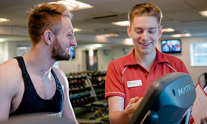 Two individuals at the gym, one using exercise equipment and the other assisting.