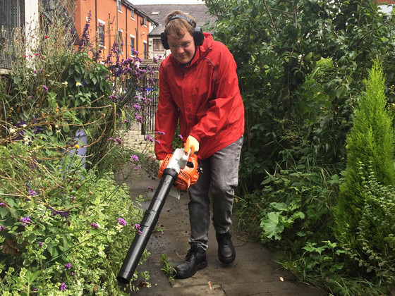 A person in a red jacket using a leaf blower in a garden.