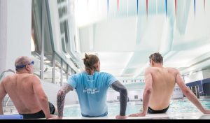 Three people sitting by an indoor swimming pool, one wearing a blue shirt with text on the back.