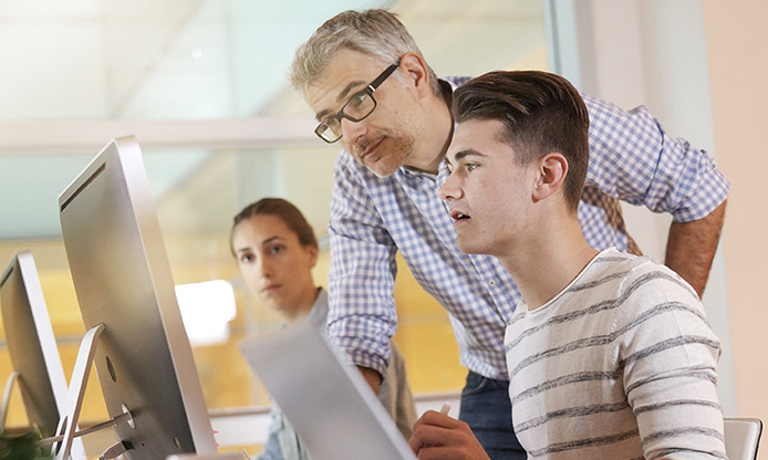 Two individuals in an office setting, one seated at a computer, the other standing.