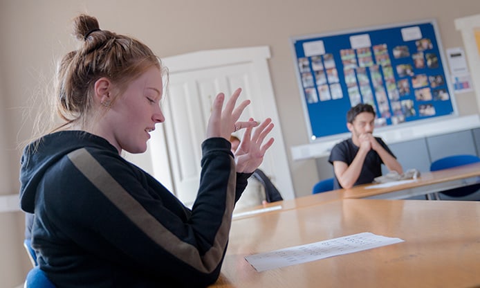 A student with raised hands appears to be speaking in a classroom setting.