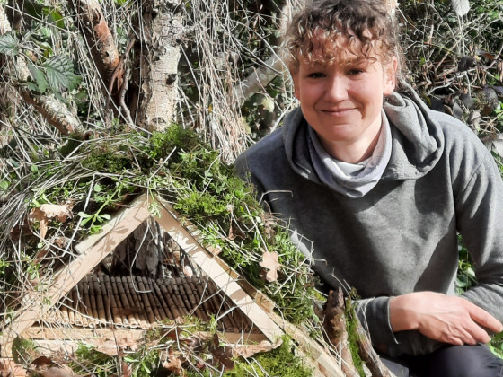 Claire next to a small triangular wooden structure covered with vegetation outdoors.