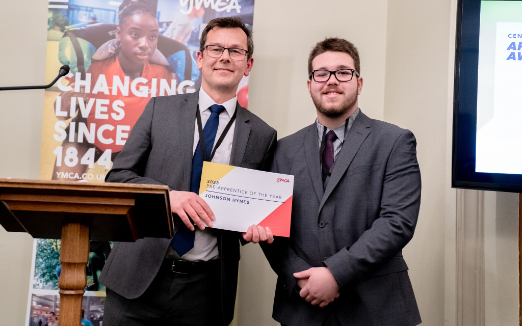 Two individuals in suits holding an 'Apprentice of the Year' certificate at a formal event.