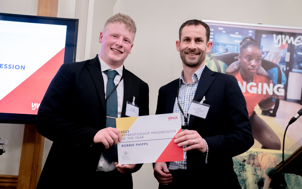 Two individuals at an event, one holding an award certificate, with a YMCA banner in the background.