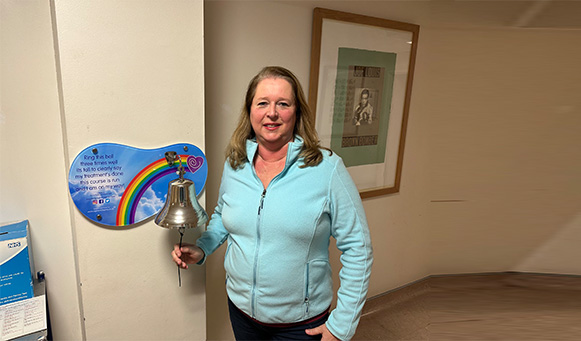 Claire standing next to a celebratory bell with rainbow plaque in a hallway.