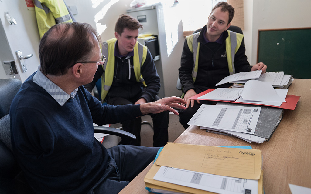 Three individuals with documents engaged in a discussion at a cluttered office table.