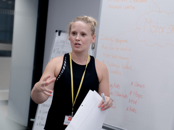 Person gesturing during a presentation with a whiteboard in the background.