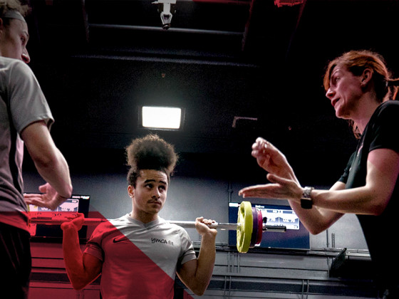 Person lifting weights in a gym with two trainers observing.