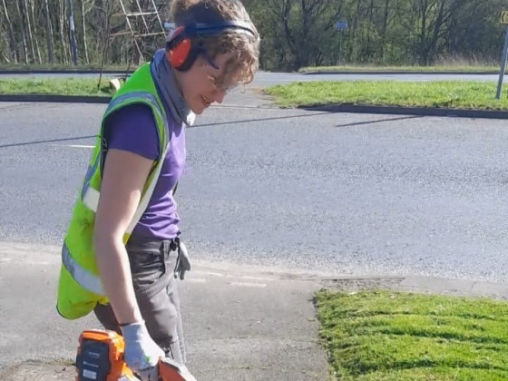 Claire wearing a hi-vis vest holding a leaf blower on a sunny day.