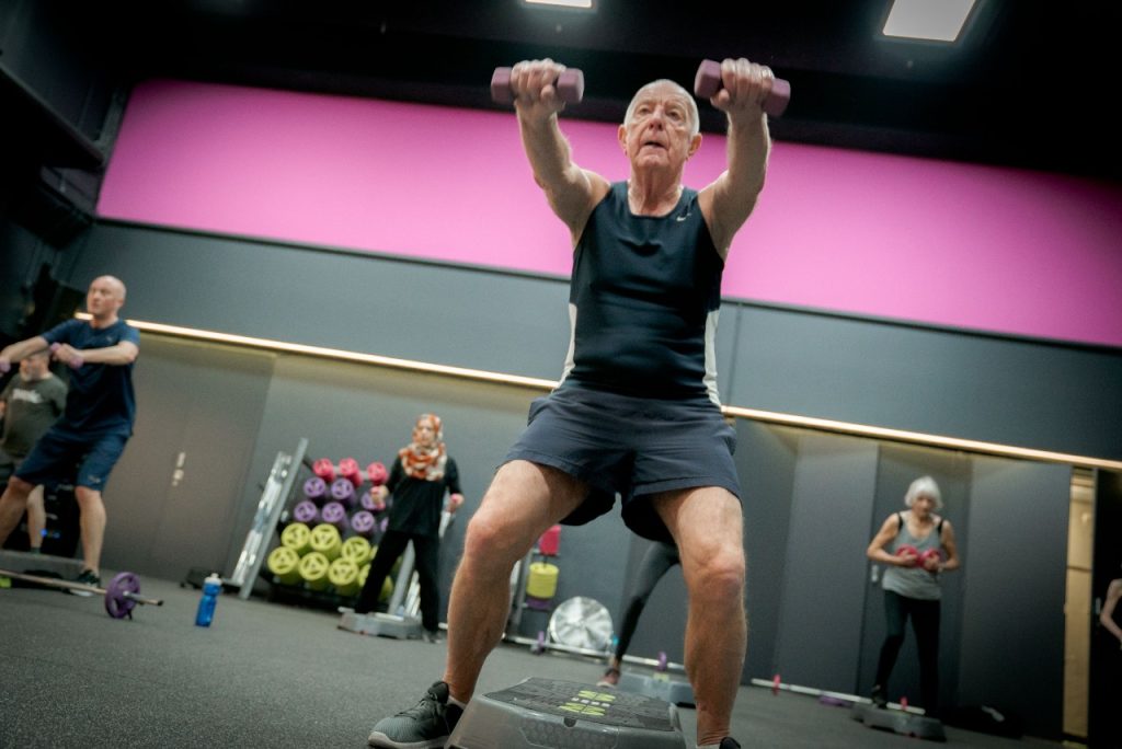 Person lifting dumbbells in a gym class with others exercising in the background.