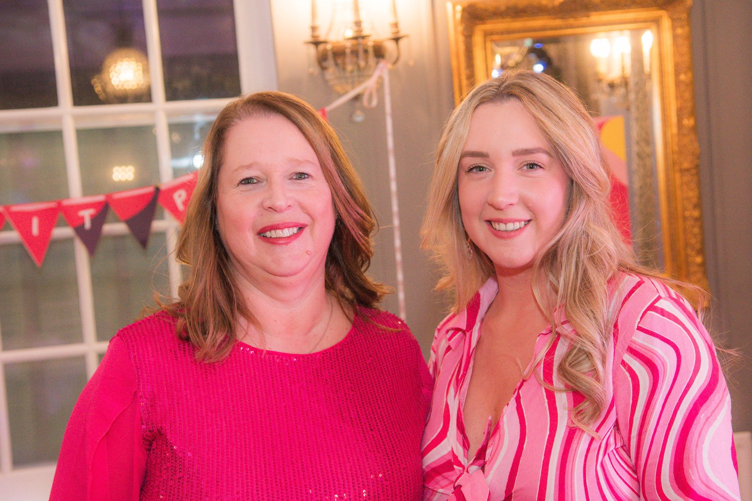 Claire and another woman in colorful attire at an indoor event with a festive banner in the background.