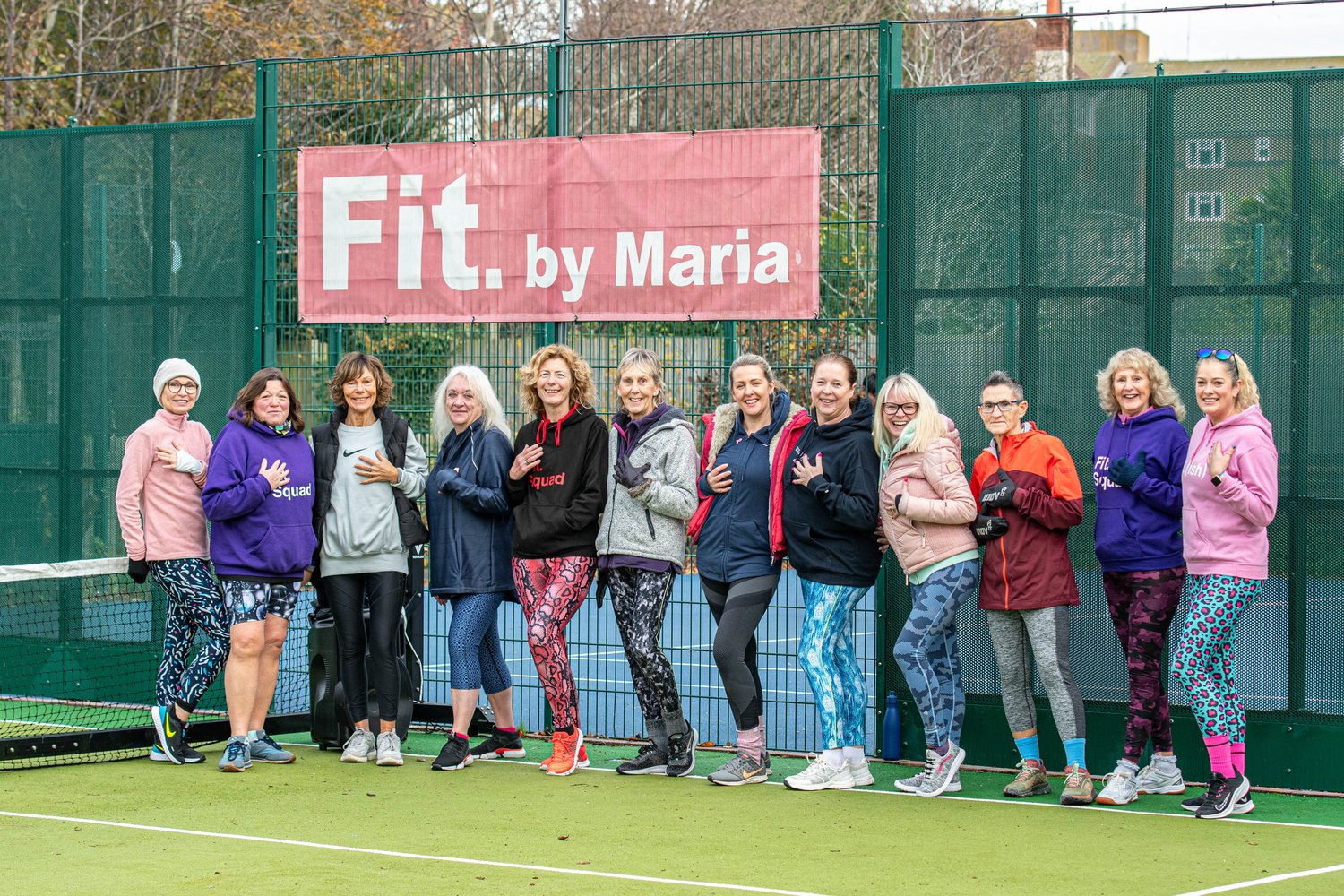 Group of people in workout gear standing in front of a "Fit. by Maria" banner at a sports court.