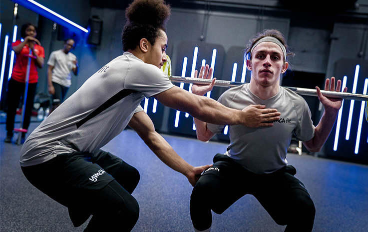 Two individuals in a gym performing a squat exercise with a barbell, surrounded by blue neon lights.