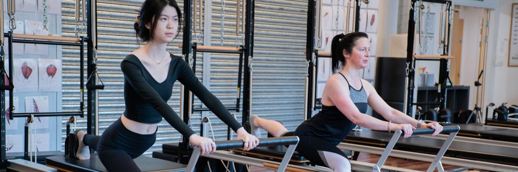 Two people exercising on pilates reformer machines in a studio.