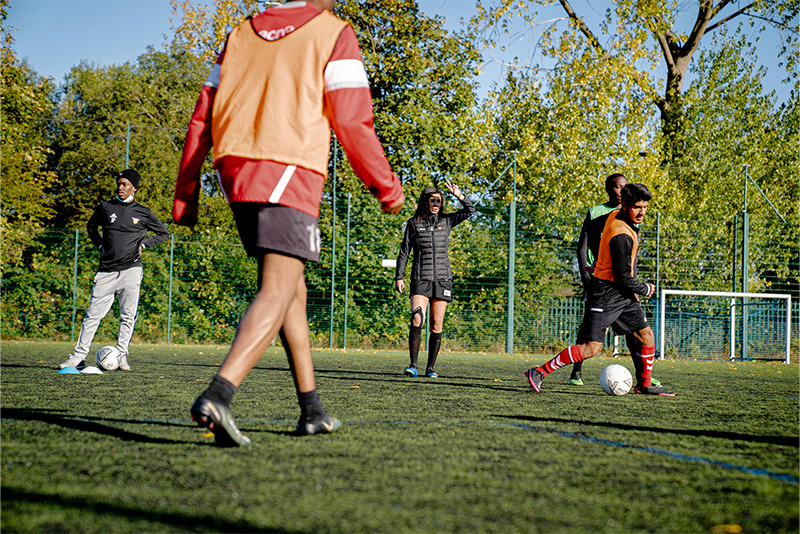 A group of young athletes in orange training vests practicing soccer on a grassy field, with goalposts and trees in the background.